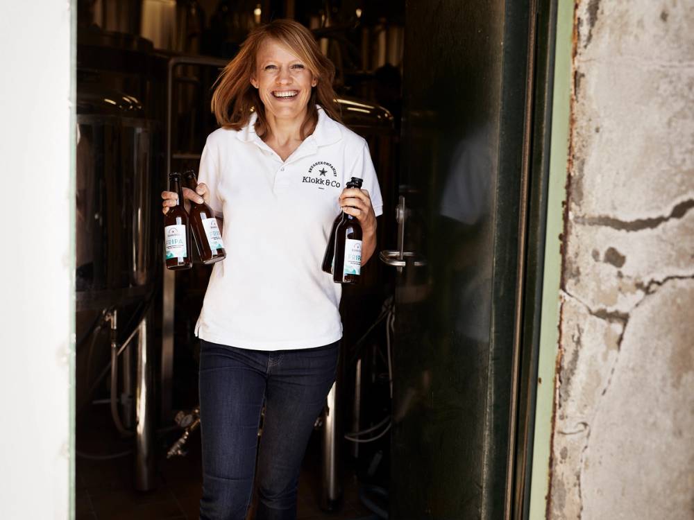 En person med skulderlangt hår som bærer to flasker med drikke, står foran en inngang til en bygning.