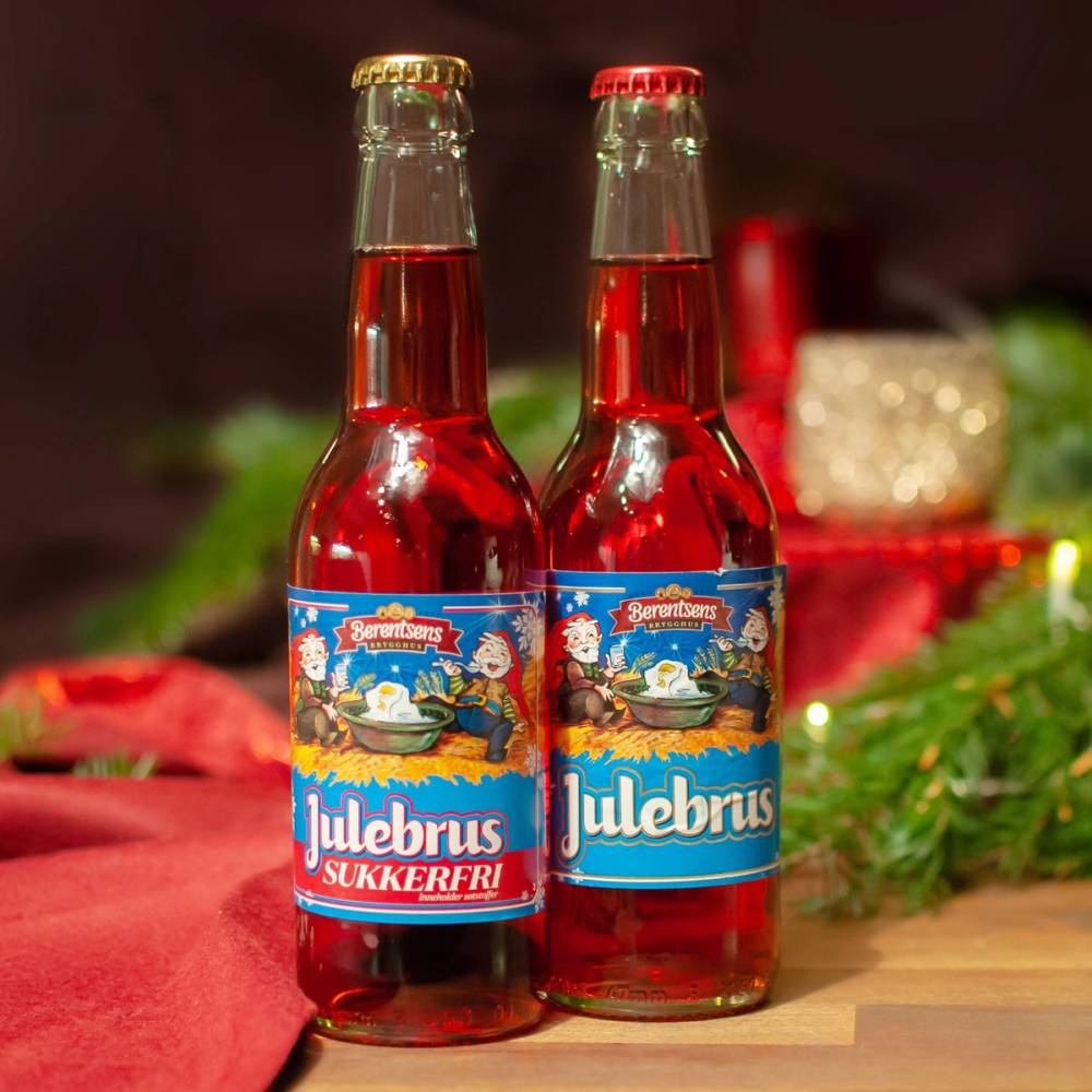 To flasker med rød julebrus med julebrus-etiketter, plassert på et bord med julepynt rundt.