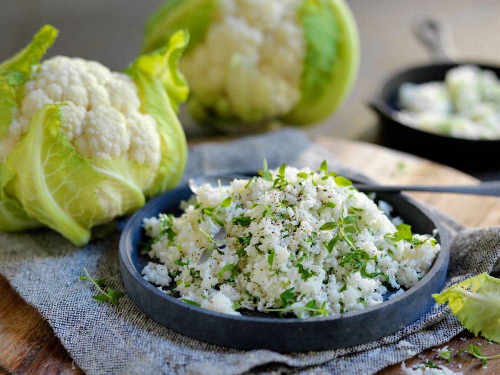 Bilde av blomkålris i en skål med blomkål og hele blomkål på siden, samt en liten svart bolle med ekstra blomkål i bakgrunnen.