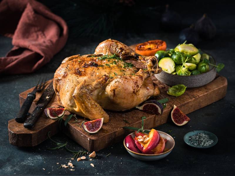 Hel stekt julekylling på et skjærebrett med fiken, grønnsaker og krydder ved siden av.