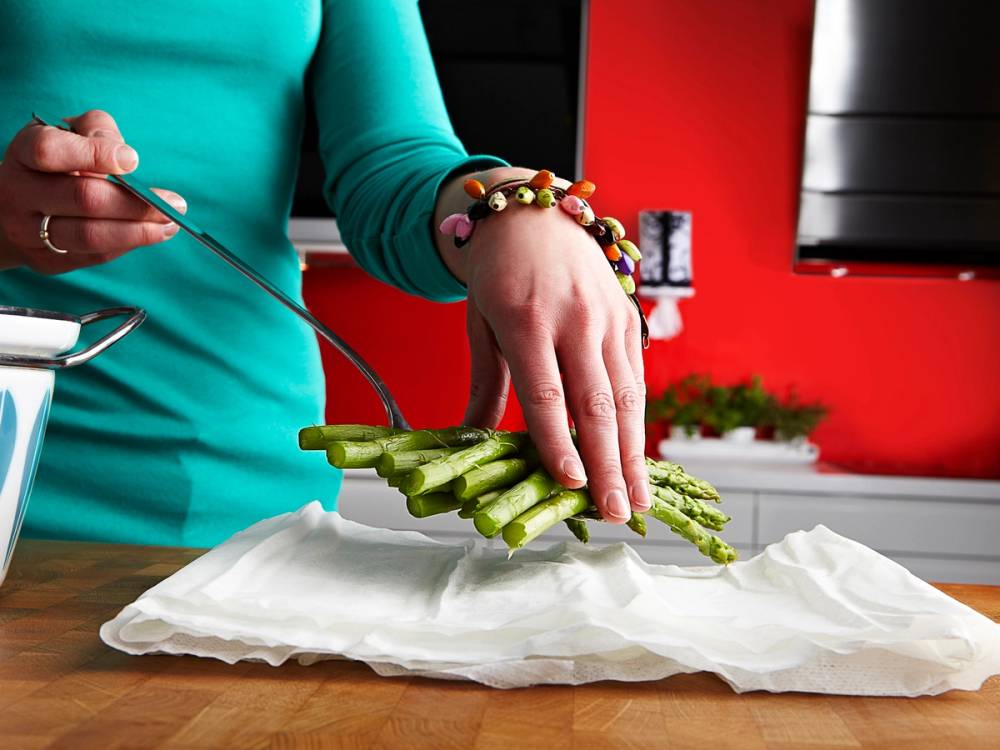 En person legger asparges på et stykke papir i et kjøkken med røde vegger, og personen har på seg en blå langermet genser og flere armbånd.