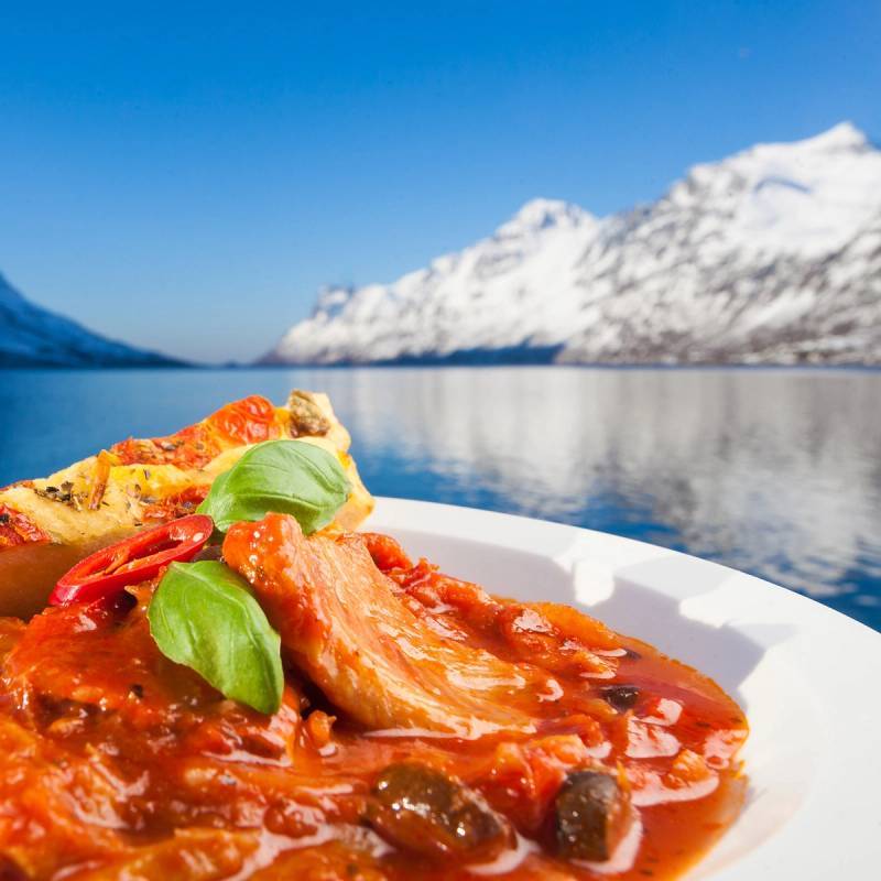 En tallerken med pizza med tomatsaus, ost, og grønne basilikumblader foran en fjord med snødekte fjell i bakgrunnen.