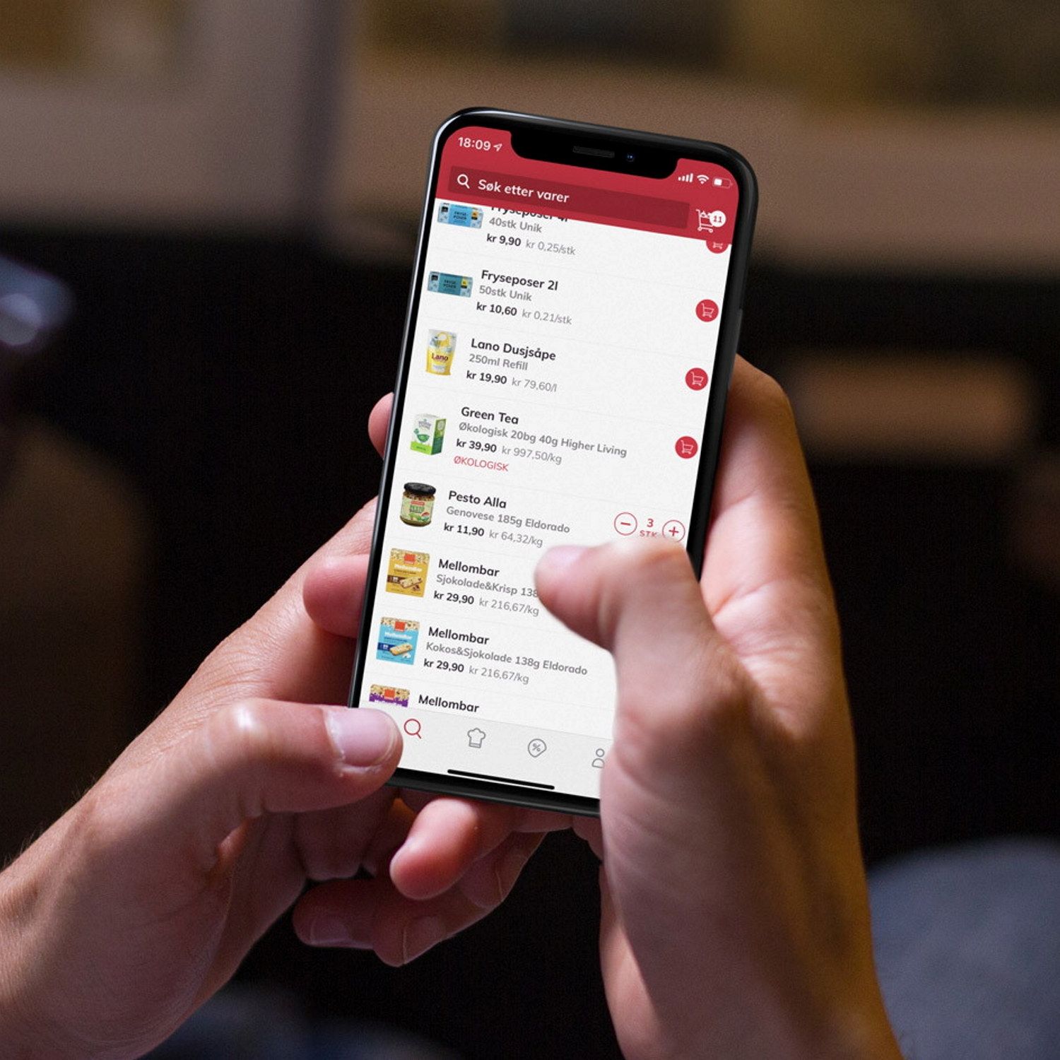 Person holder en mobil med en app som viser en liste over ulike produkter eller tjenester, med en rød toppmeny og ikoner nederst på skjermen.