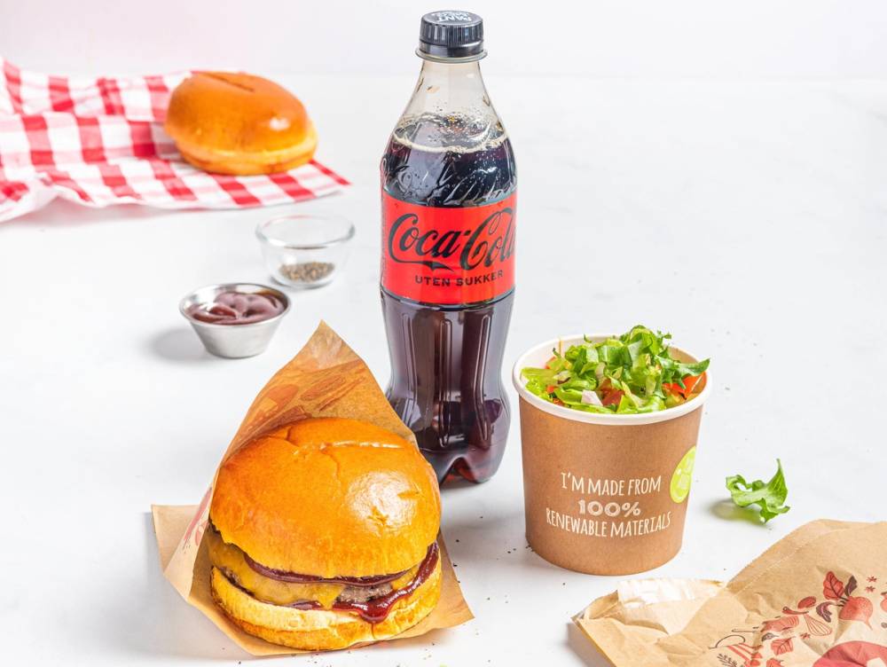 En burger med pommes frites, en flaske Coca-Cola og en salat i en papirskål på et hvitt bord.