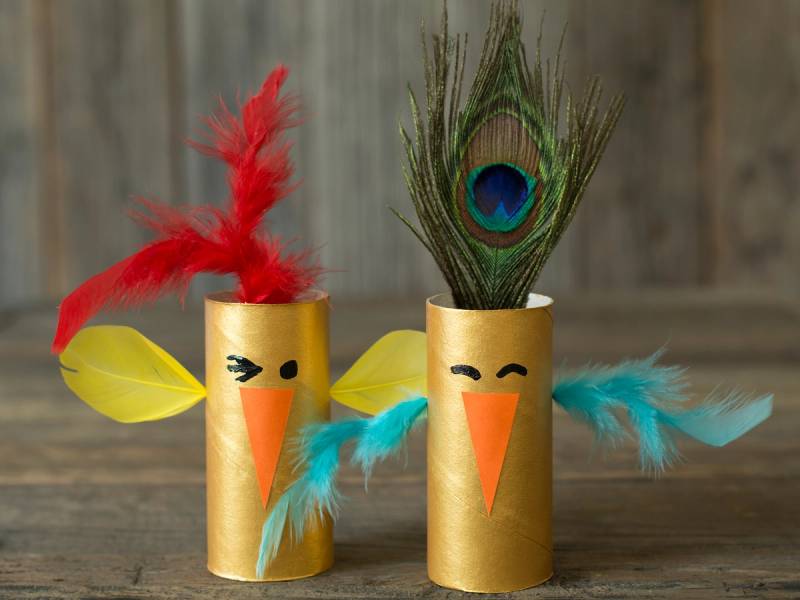 To påskepynt laget av tomme toalettpapirruller med fjær og papirfigurer som ser ut som kyllinger, plassert på en treflate mot en trevegg.