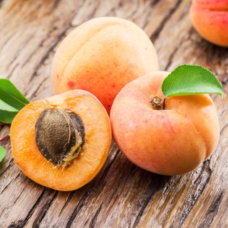 To ferske aprikoser, en hel og en halvert med steinen synlig, ligger på en rustikk treflate med en grønn blad ved siden av den hele aprikosen.