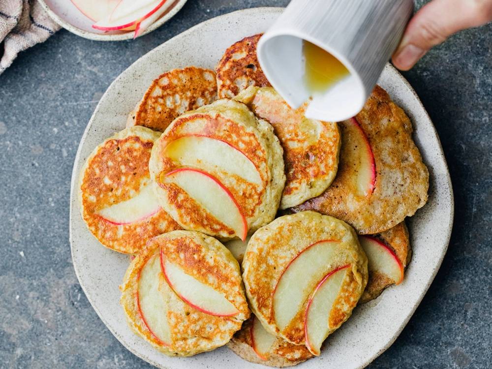 Pannekaker med skiver av eple på en tallerken, med en hånd som heller lønnesirup over dem