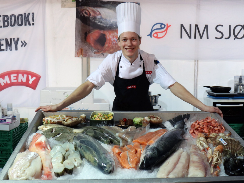 En kok i hvit uniform og kokkehatt står bak en fiskebuffet med ulike typer rå sjømat på en butikkdisk.