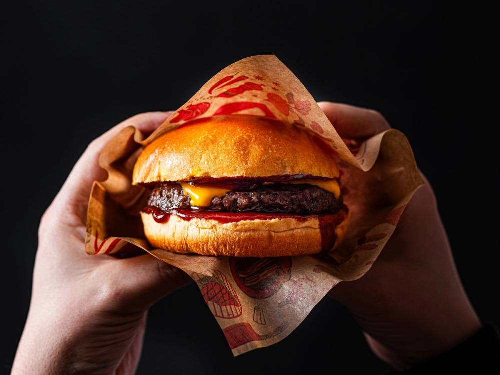 En person holder en burger med kjøtt, ost og brød i hånden, med en mørk bakgrunn.