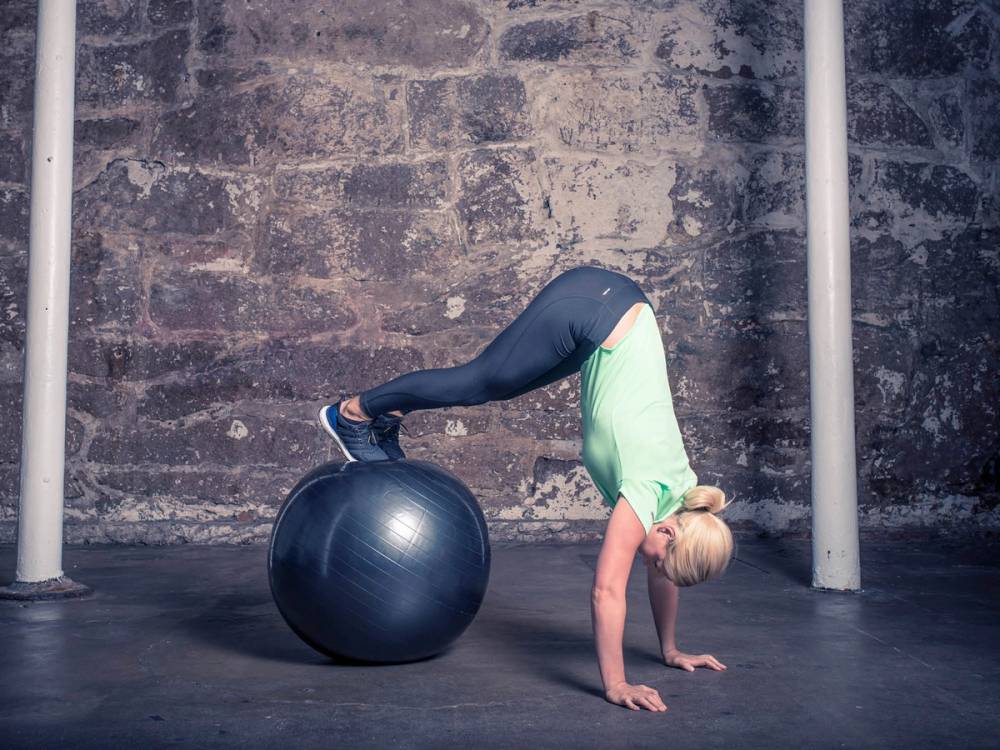 En kvinne i treningsklær som står med hendene på gulvet og føttene på en stor treningsball, i et treningsrom med murvegg.