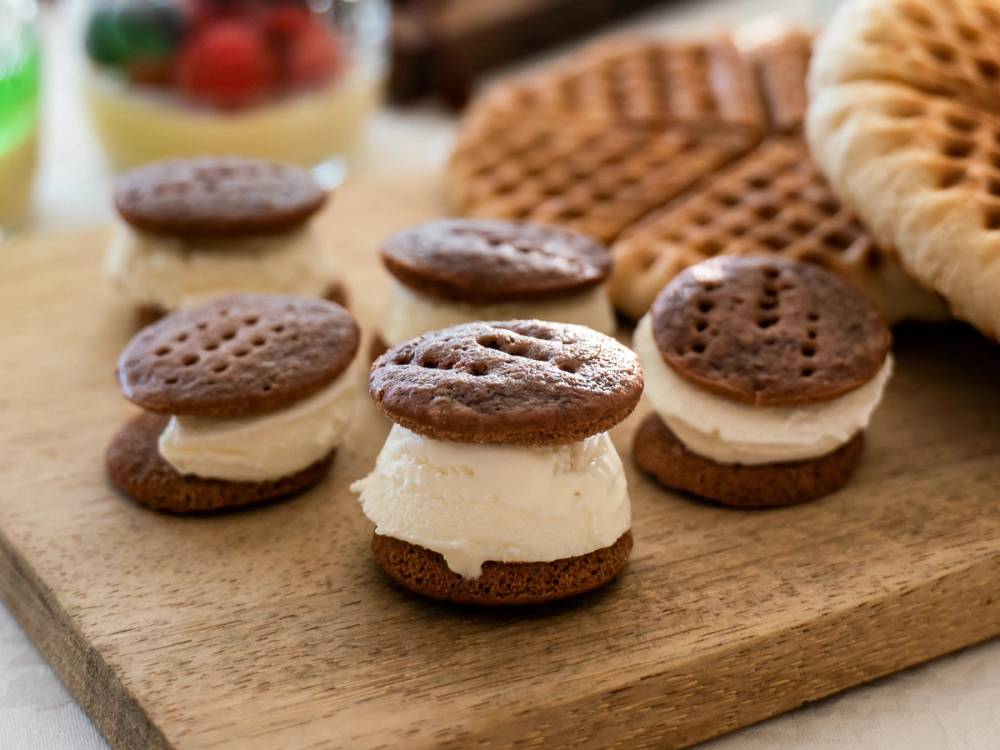 Småkaker med sjokolade og krem på et trebrett, omgitt av andre kaker og frukt i bakgrunnen.