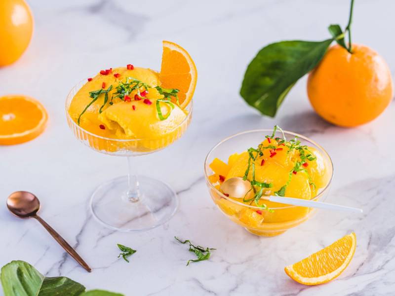 To glass med appelsinsorbet pyntet med sitronskiver og små røde bær, omgitt av hele appelsiner og appelsinbiter på en hvit bakgrunn.