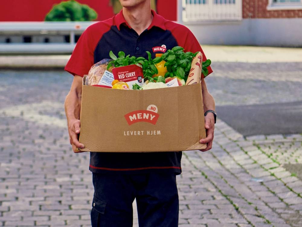 Person som holder en stor eske med mat fra Meny, klar til levering hjemme, med en bakgrunn av gate og fortau.