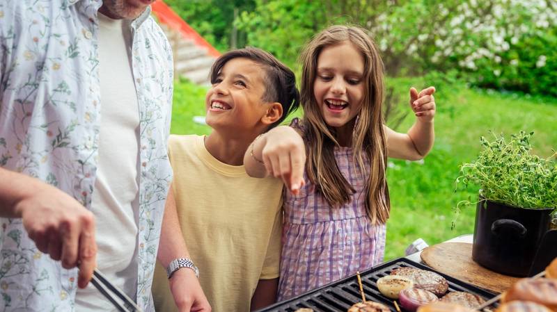 To barn som står ved en grill med mat, mens en voksen hjelper til, i en grønn hage.