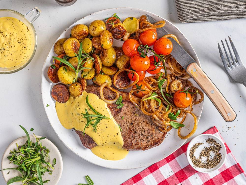 Stekt biff med bearnaisesaus, småpoteter, cherrytomater og løk på en hvit tallerken, omgitt av urter og krydder.
