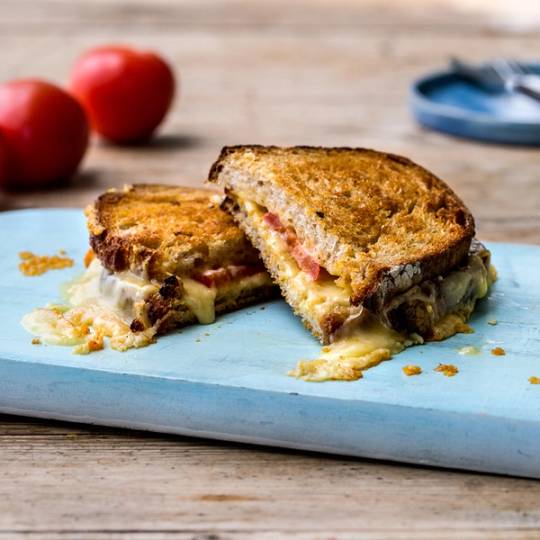 Et ostesmørbrød med norsk alpeost og sennep som er delt i to, på et hvitt skjærebrett med tomater i bakgrunnen.