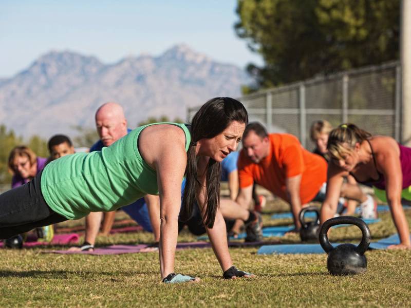 En gruppe mennesker deltar i en utendørs treningsøkt med push-ups og kettlebell-øvelser på gresset, med fjell i bakgrunnen.