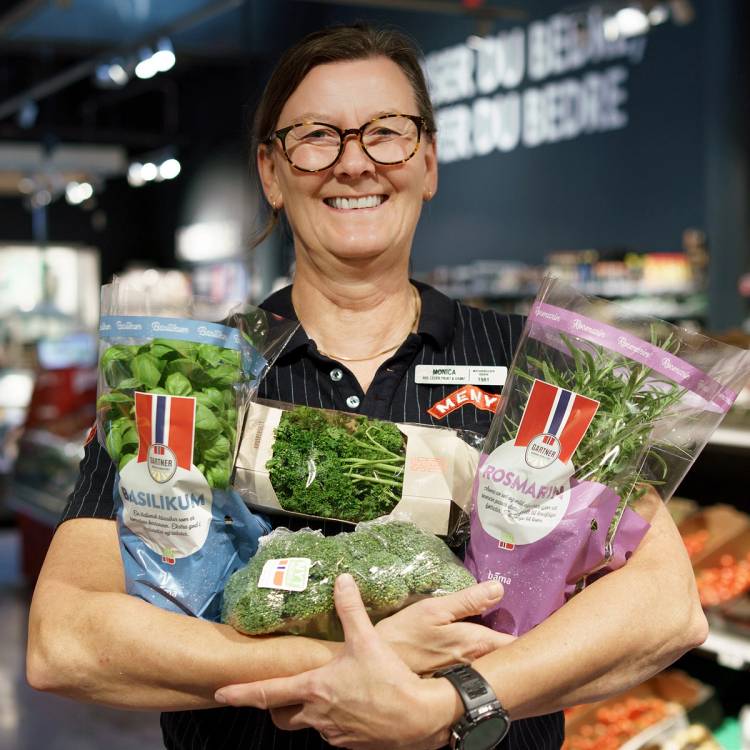 En person i butikk holder flere pakker med grønnsaker og urter foran seg, med butikkhyller i bakgrunnen.
