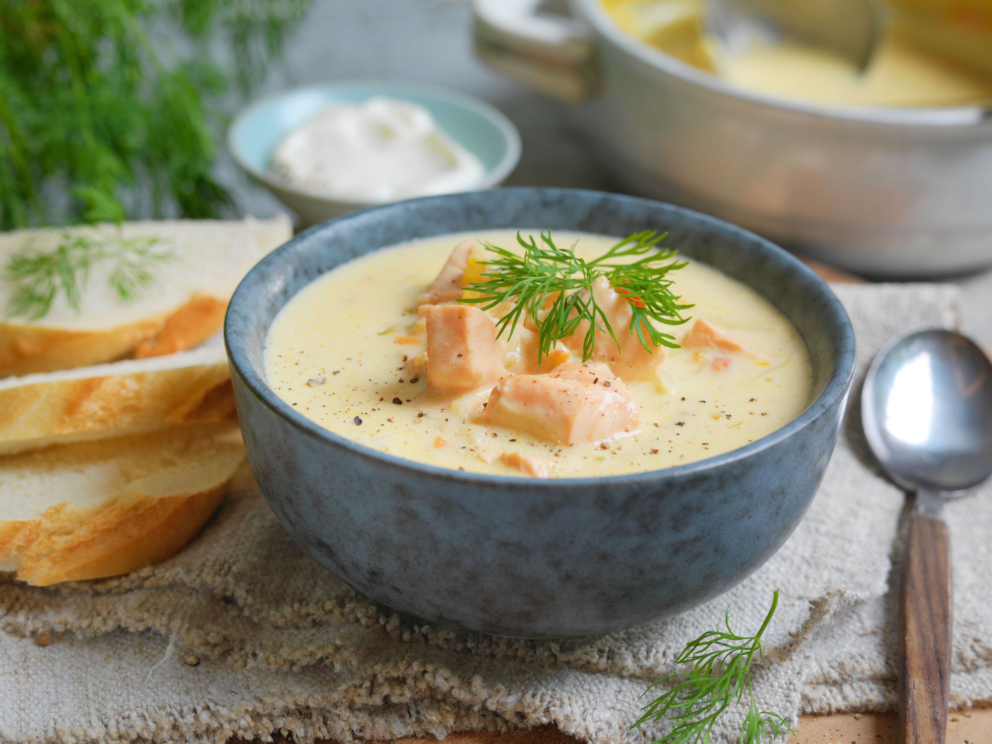 Fiskesuppe servert i en blå bolle med fersk dill på toppen, omgitt av brødskiver og tilbehør.