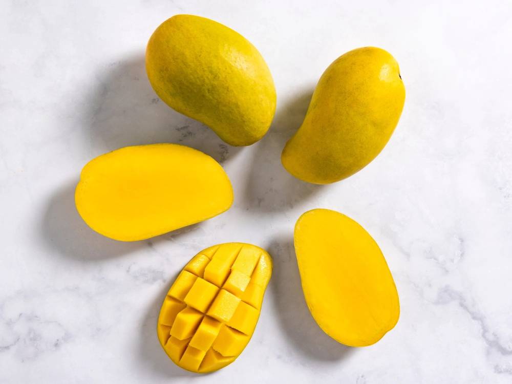 Fire gule mangoer, hvorav en er skåret opp i terninger, lagt på en hvit overflate.