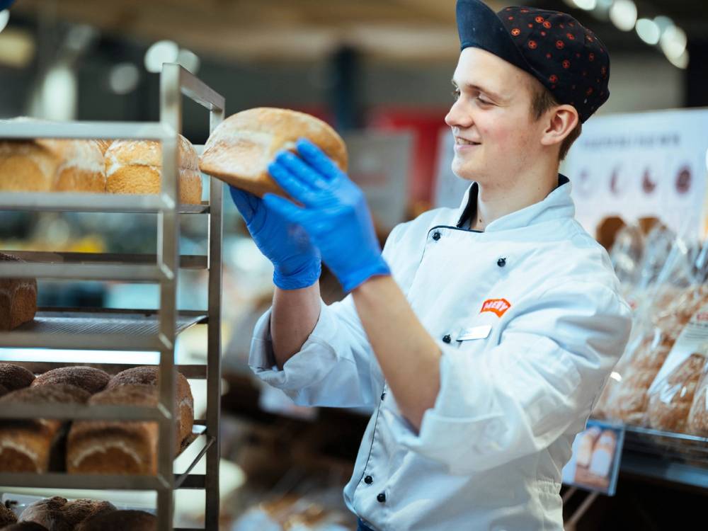 En baker i hvit uniform og blå hansker tar ut et brød fra en ovn i et bakeri.