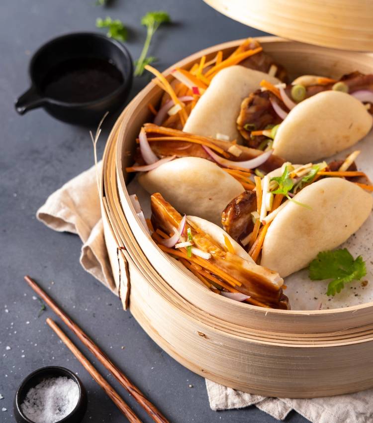 Steambuns med fyll av kjøtt og grønnsaker i en bambusdampkurv, med spisepinner og små skåler ved siden av.