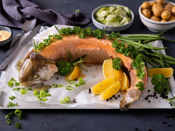 Hel, bredde av en stekt eller bakt hel fisk på et serveringsfat med sitronbåter og grønne urter, omgitt av ulike tilbehør og grønnsaker.