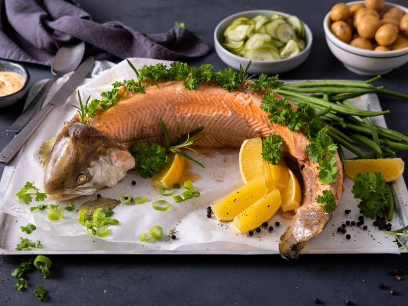 Hel, bredde av en stekt eller bakt hel fisk på et serveringsfat med sitronbåter og grønne urter, omgitt av ulike tilbehør og grønnsaker.