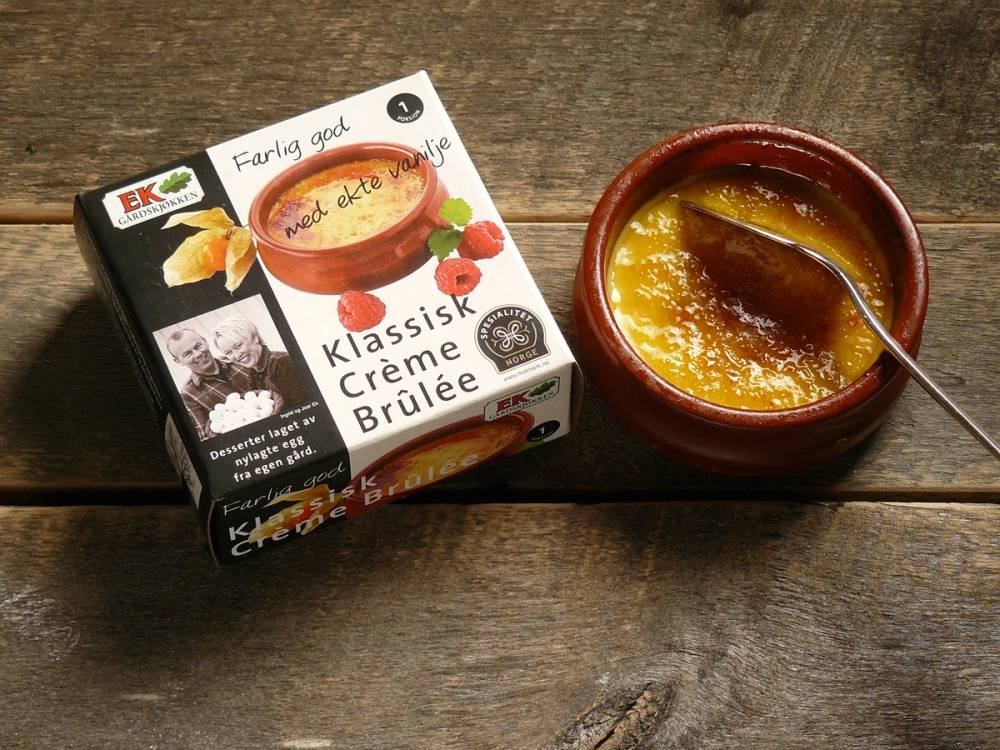 En kopp med brunt innhold og en skje ved siden av, plassert på et trebord, med en eske med EKs klassiske crème brûlée ved siden av.