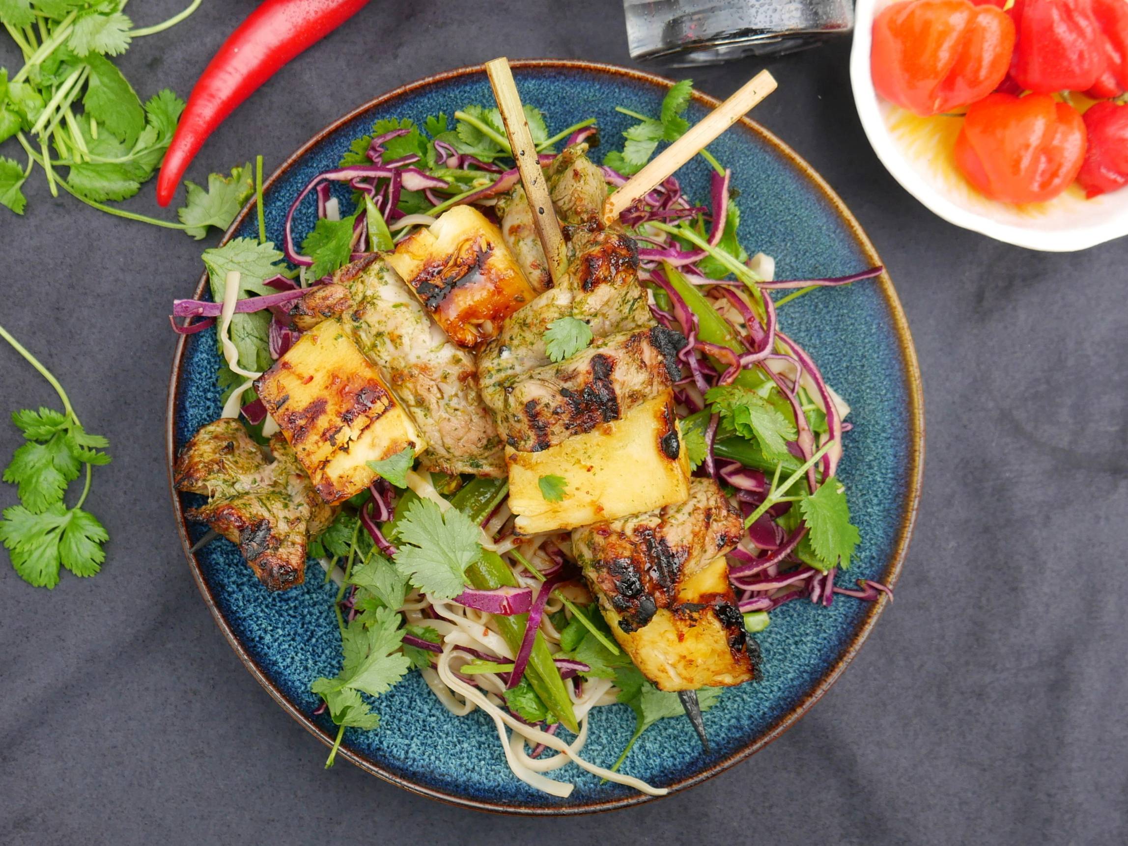 Grillspyd med kjøtt og grønnsaker på en blå tallerken, omgitt av friske urter og tomater i en liten skål, på en mørk bakgrunn.