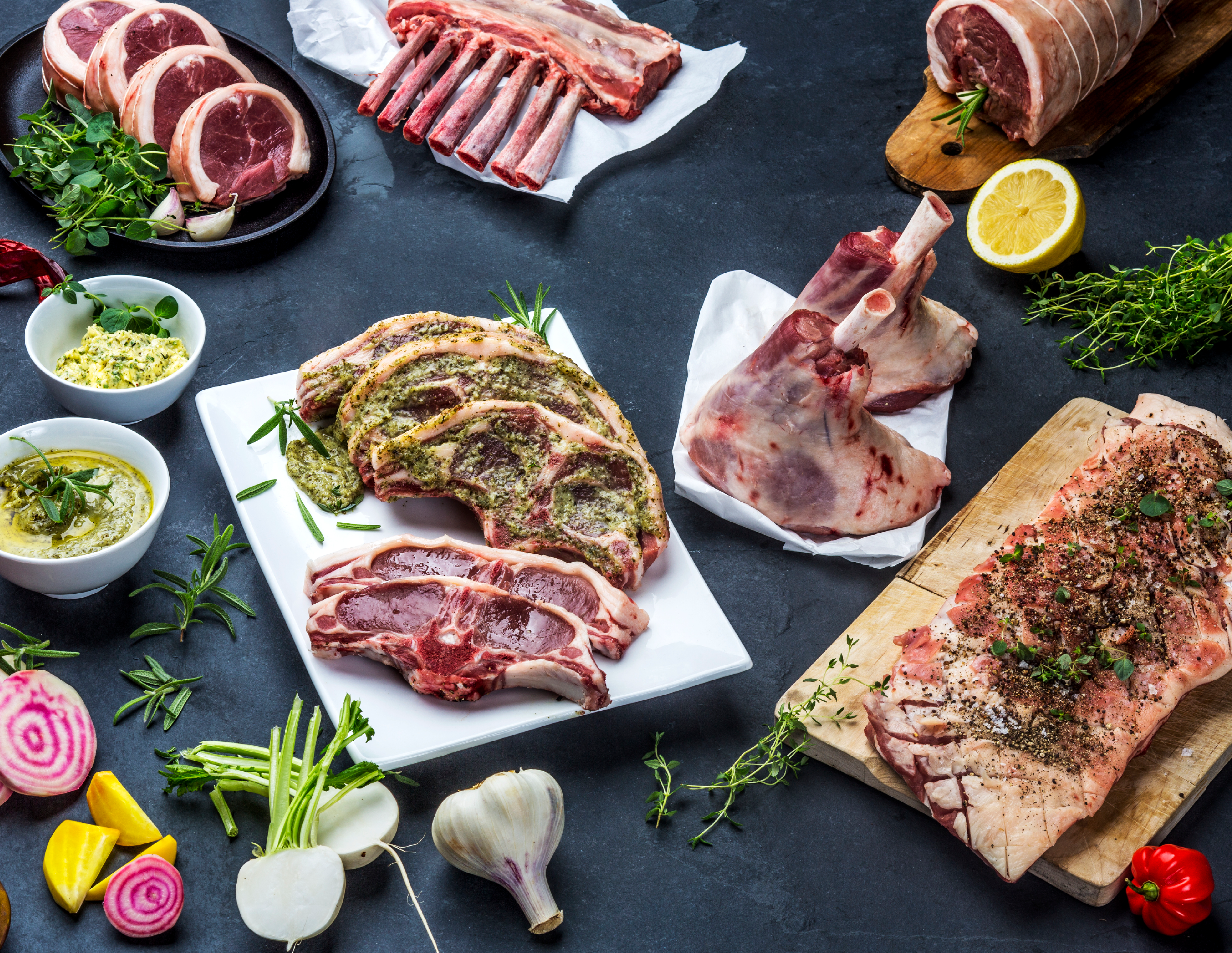 Ulike typer kjøtt og urter på et kjøkkenbord, klare til å bli tilberedt, med noen små grønnsaker og krydder rundt.