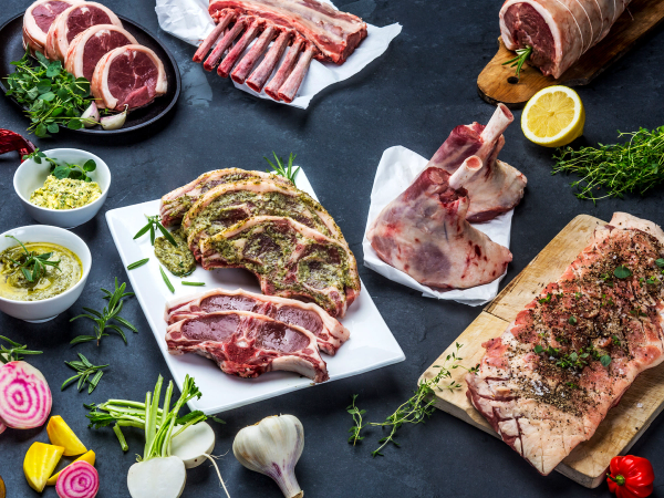 Ulike typer kjøtt og urter på et kjøkkenbord, klare til å bli tilberedt, med noen små grønnsaker og krydder rundt.