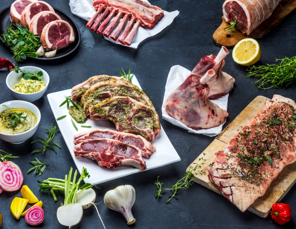 Ulike typer kjøtt og urter på et kjøkkenbord, klare til å bli tilberedt, med noen små grønnsaker og krydder rundt.