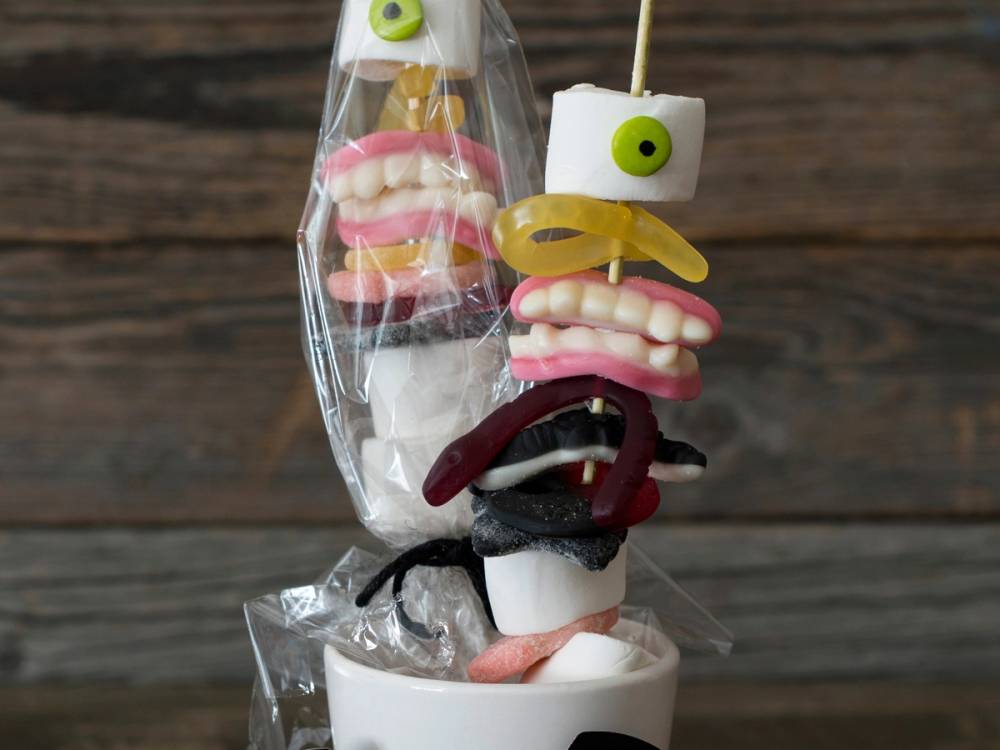 Små kaker på grillpinner med ulike farger og dekorasjoner, pakket inn i plast, på en treoverflate.