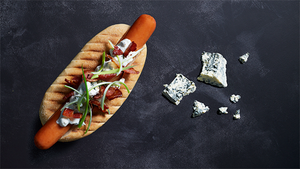 En pølse med ost og urter på en trepinne, med blåmuggostbiter ved siden av på en svart bakgrunn.