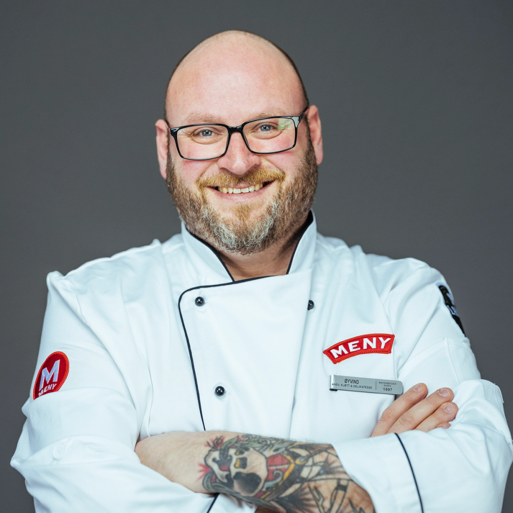 En person i hvit kokkeflate med armer korslagt, med tatoveringer på armen, mot en grå bakgrunn.