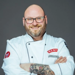 En person i hvit kokkeflate med armer korslagt, med tatoveringer på armen, mot en grå bakgrunn.