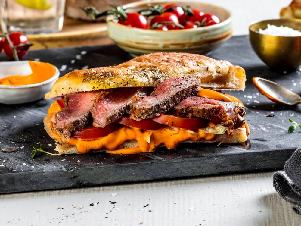 Et smørbrød med skiver av lam, tomat og salat på en svart skjærefjøl, omgitt av små skåler med saus og tilbehør.