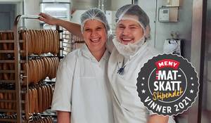 To personer i hvite kokkerklær står i en butikk med metervare, og en av dem peker mot hyller med ruller av stoff eller papir, med en logo for Meny Mat Skatt i hjørnet.