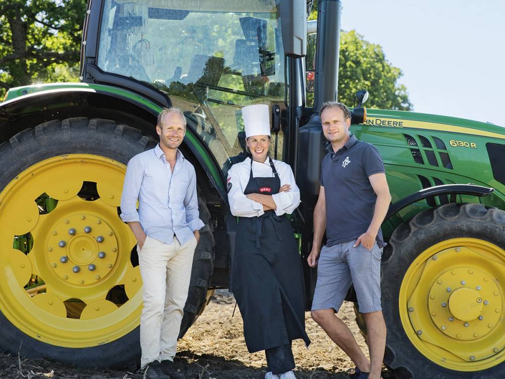 Tre personer står foran en stor grønn og gul traktor på en jordete vei, med trær i bakgrunnen.
