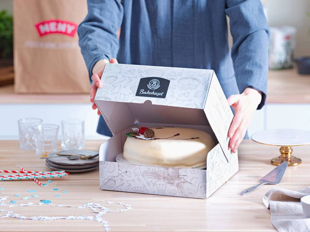 En person åpner en hvit kakeeske med en kake inni på et kjøkkenbord, omgitt av kakeutstyr og pynt.