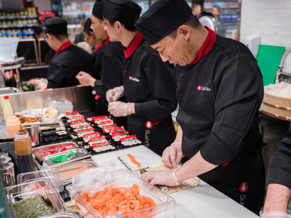 Kjøkkensjefer i svart uniform og rødt skjerf jobber med matlaging i en butikk eller restaurant, med ulike ingredienser og utstyr på benken.