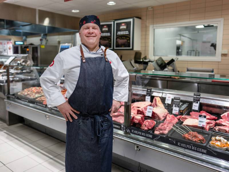 En person i butikkuniform står foran kjøledisken med kjøttprodukter i en MENY-butikk, med fokus på personens klær og butikkmiljøet.