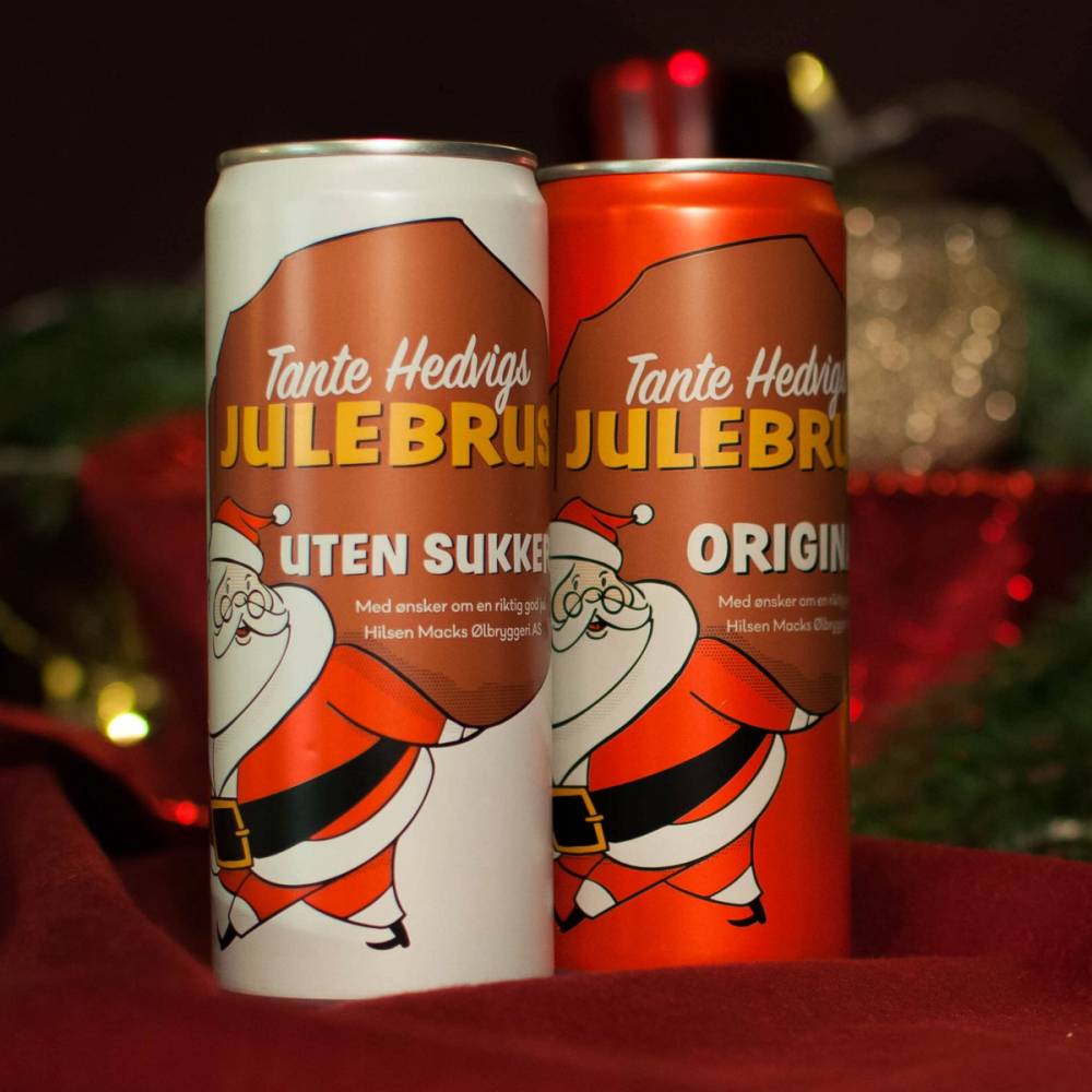 To bokser med julebrus fra Tante Hedvig, en med hvit etikett og en med oransje etikett, plassert på et bord med julepynt i bakgrunnen.