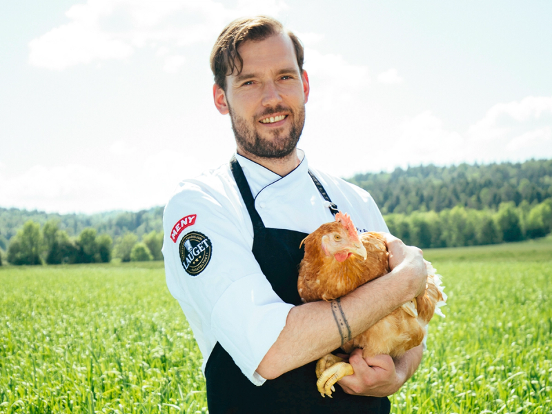 En person i kokkeforkledning holder en kylling i armene ute i en grønn eng med trær i bakgrunnen.