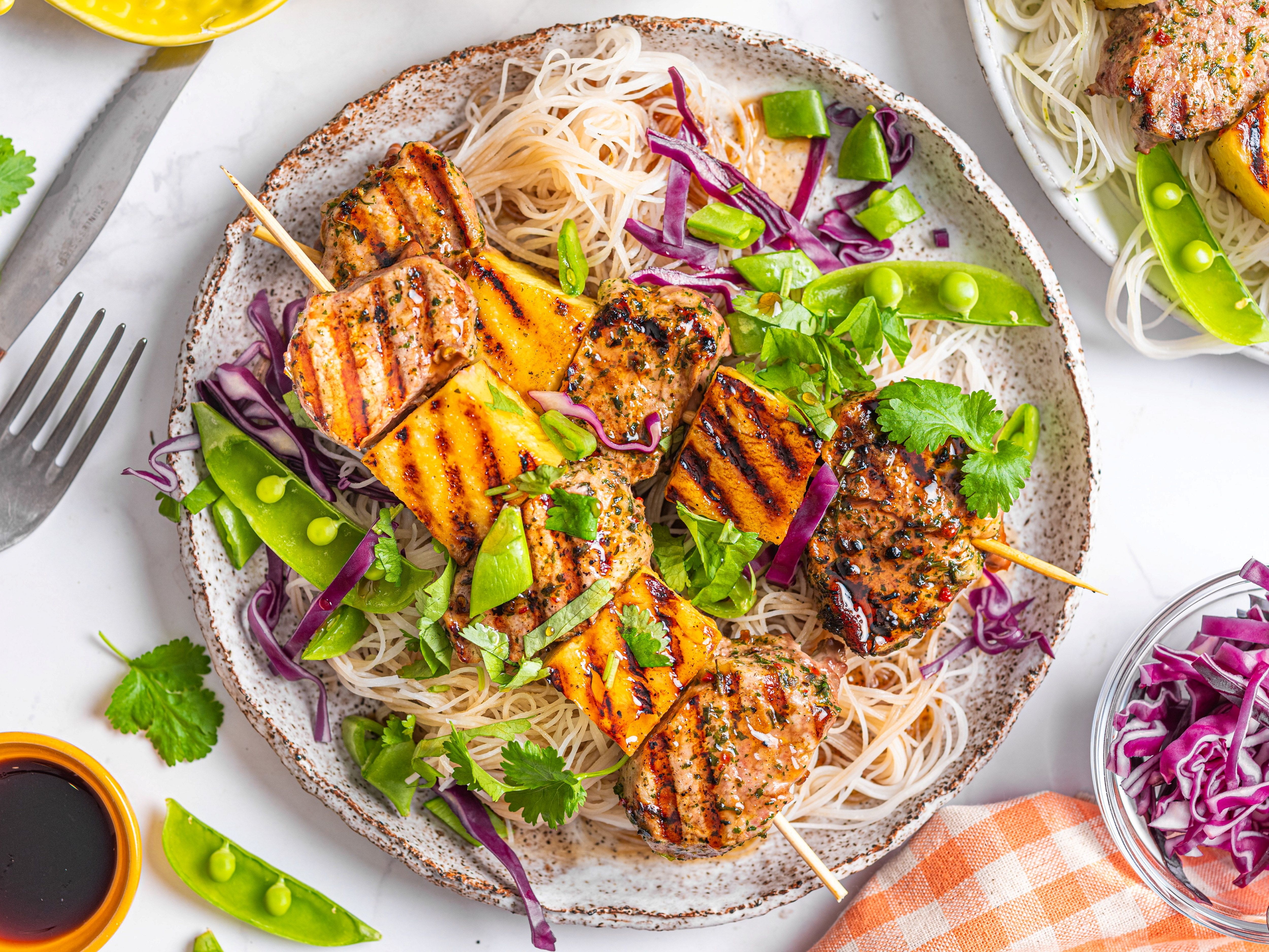 Thai salat med grillspyd av svinefilet, servert med risnudler og grønnsaker som paprika og reddiker, på en hvit tallerken.
