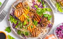 Thai salat med grillspyd av svinefilet, servert med risnudler og grønnsaker som paprika og reddiker, på en hvit tallerken.