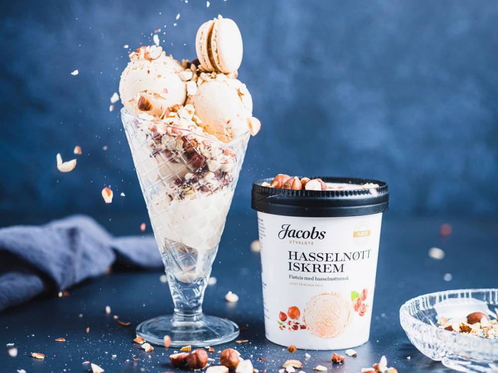 Iskrem i en glass med flere kuler og en boks med Jacobs Hasselnøtt iskrem ved siden av, mot en blå bakgrunn.
