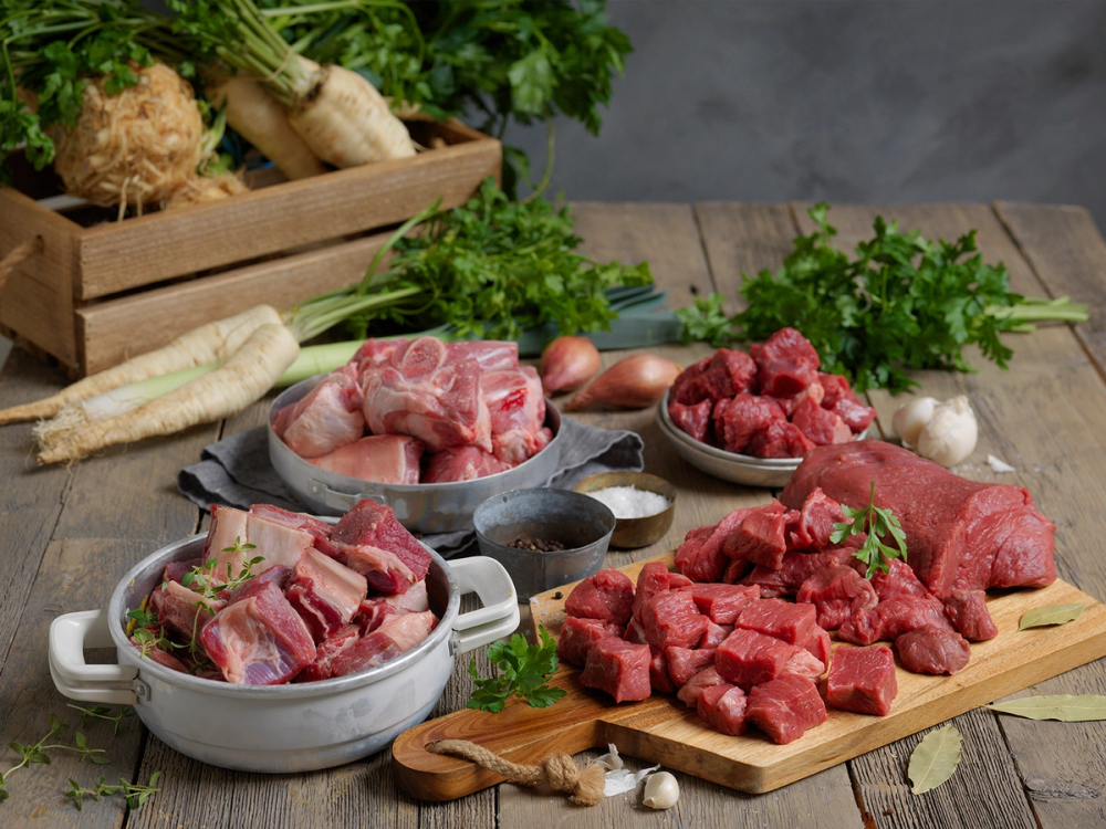 Ulike typer av suppekjøtt og kjøttstykker på en kjøkkenbenk med grønnsaker og krydder rundt.