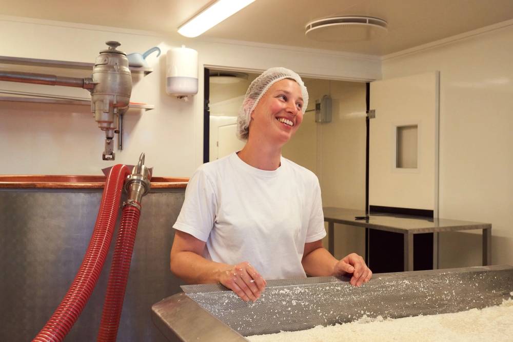 En person i hvit kjeledress står bak en stor, flat overflate i et ysteri, med utstyr og rør på veggen i bakgrunnen.